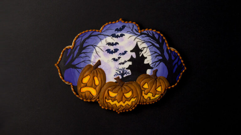 Spooky Jack O'Lantern Cookie for Halloween - a plaque shaped cookie decorated with a full moon, a silhouette of a haunted house on a hill surrounded by spooky trees and bats. 3 glowing jack o'lanterns are in the foreground. There is an orange bead border around the edge of the cookie.