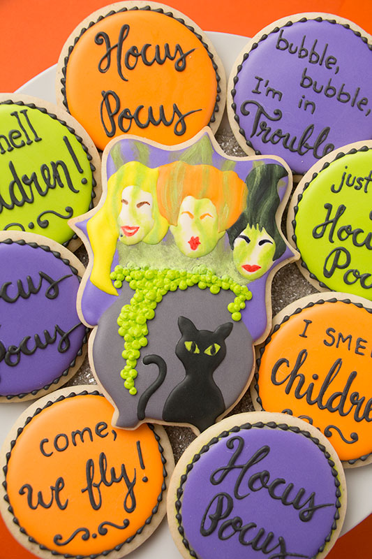 Hocus Pocus cookies - a set of cookies decorated in bright orange, green, and purple royal icing. There is a plaque shaped cookie with the Sanderson sisters' faces in the middle. The faces are above a bubbling cauldron with green mist rising from it. There is a black cat in front of the cauldron. There are round cookies surrounding the plaque cookie with quotes from the move piped on them. The cookies are on a white platter on an orange background.