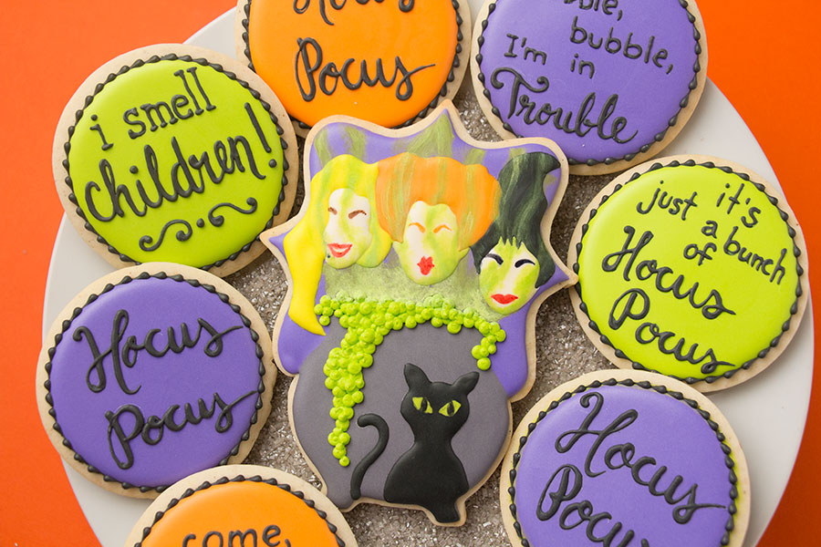 Hocus Pocus cookies - a set of cookies decorated in bright orange, green, and purple royal icing. There is a plaque shaped cookie with the Sanderson sisters' faces in the middle. The faces are above a bubbling cauldron with green mist rising from it. There is a black cat in front of the cauldron. There are round cookies surrounding the plaque cookie with quotes from the move piped on them. The cookies are on a white platter on an orange background.