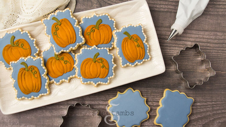 Decorated pumpkin cookies - a set of decorated cookies in ornate plaque shapes decorated with royal icing pumpkins on a blue background. The cookies are on a rectangle platter textured with a cable knit pattern. The platter is on a wood background. there is a piece of lace fabric above the platter, two cookie cutters and two iced cookies below the platter, and a decorating bag next to the platter.