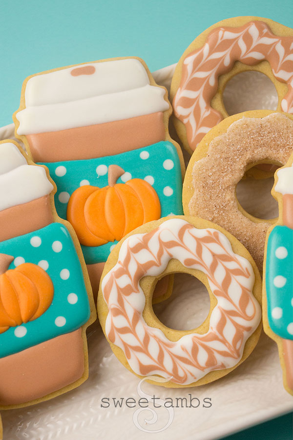 Pumpkin Spice Coffee and Donut Cookies - Cookies decorated to look like paper coffee cups and iced donuts. The coffee cups are light brown with white lids and have a teal sleeve with white polka dots. There are pumpkins on the coffee cup sleeves. Some of the donuts are decorated with a herringbone design in white and light brown icing. Some of the donuts are decorated with plain icing and dipped in a mixture of cinnamon and sugar. The cookies are on a platter with a cable knit pattern. The platter is on a teal background.