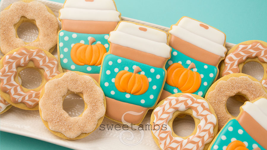 Pumpkin Spice Coffee and Donut Cookies - Cookies decorated to look like paper coffee cups and iced donuts. The coffee cups are light brown with white lids and have a teal sleeve with white polka dots. There are pumpkins on the coffee cup sleeves. Some of the donuts are decorated with a herringbone design in white and light brown icing. Some of the donuts are decorated with plain icing and dipped in a mixture of cinnamon and sugar. The cookies are on a platter with a cable knit pattern. The platter is on a teal background.