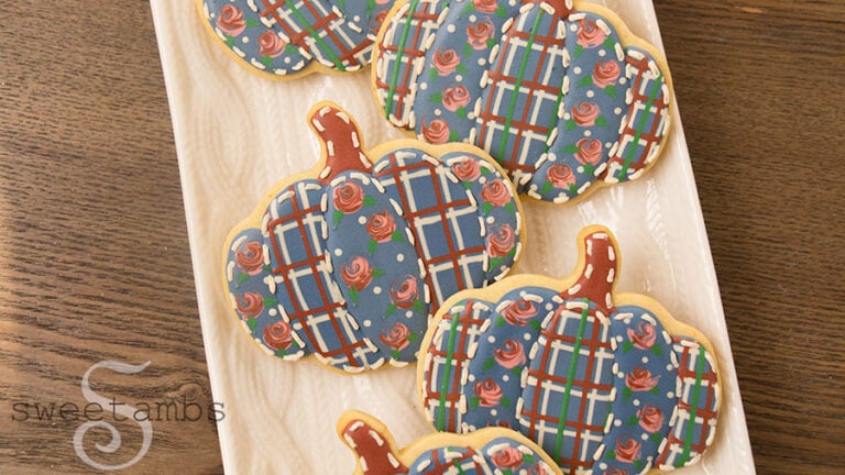 Quilted pumpkin cookies with wet-on-wet royal icing roses. These pumpkin shaped cookies are decorated with royal icing to look like they are quilted with plaid and rose patterned fabric. The cookies are on a platter with a cable knit design on a wooden backdrop.