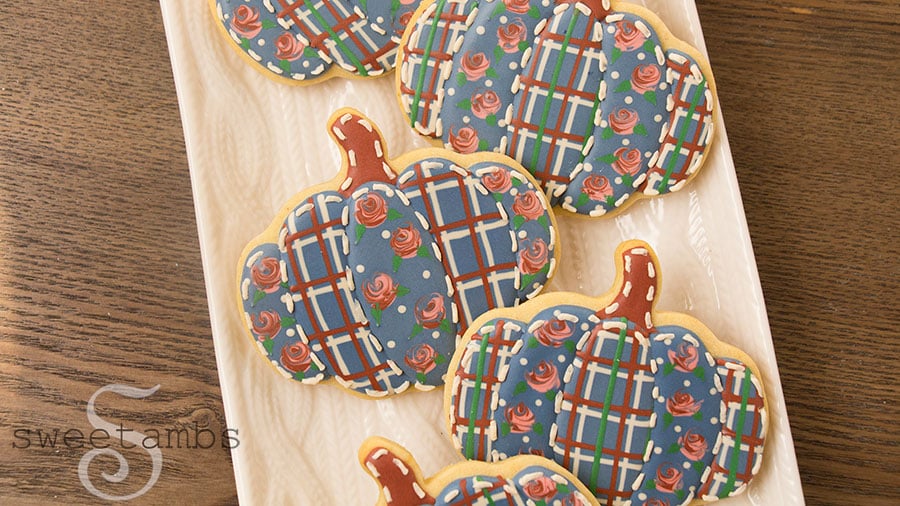 Quilted pumpkin cookies with wet-on-wet royal icing roses. These pumpkin shaped cookies are decorated with royal icing to look like they are quilted with plaid and rose patterned fabric. The cookies are on a platter with a cable knit design on a wooden backdrop.