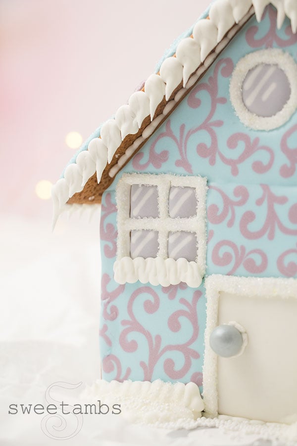 A close up of A gingerbread house decorated with royal icing. The house features a pastel blue and purple filigree design around the windows of the house. The roof is decorated with a scalloped pattern and covered in sparkling sugar. The windows of the house are sprinkled with sparkling sugar. There are royal icing icicles hanging off the edge of the roof. There is a pink bokeh background with twinkling lights.