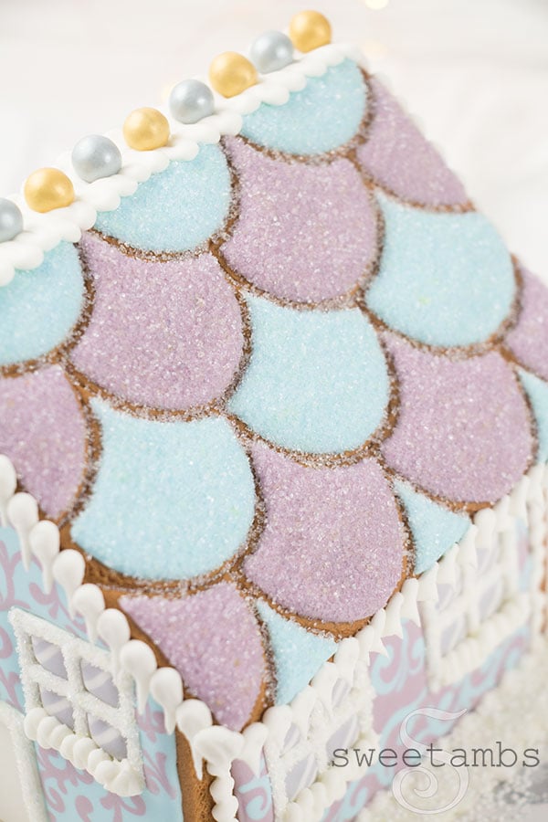 A close up of the roof of the gingerbread house. The roof is decorated with pastel blue and purple royal icing and covered with sparkling sugar. There are royal icing icicles hanging off the edges of the roof. There are gold and silver sixlets lining the top of the roof.