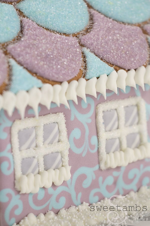 A close up of A gingerbread house decorated with royal icing. The house features a pastel blue and purple filigree design around the windows of the house. The roof is decorated with a scalloped pattern and covered in sparkling sugar. The windows of the house are sprinkled with sparkling sugar.