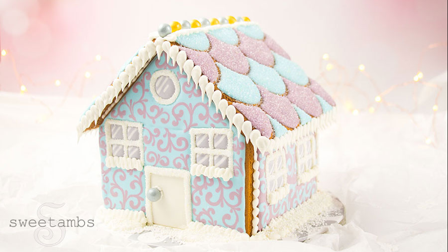 A gingerbread house decorated with royal icing. The house features a pastel blue and purple filigree design on the front and sides. The roof is decorated with a scalloped pattern and covered in sparkling sugar. The windows and doors of the house are sprinkled with sparkling sugar. The house is sitting on a white base and there is a pink bokeh background with twinkling lights.