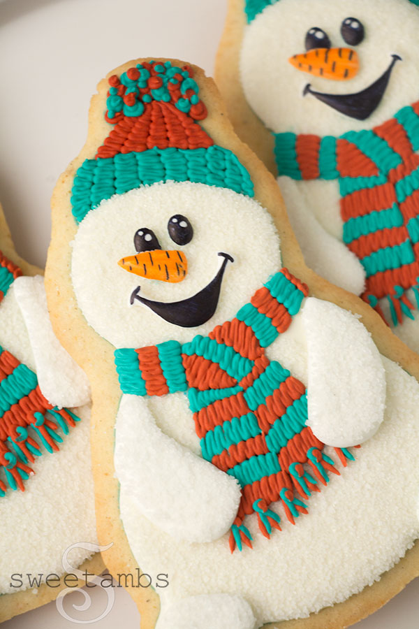 Close up of snowman cookies on a white plate on a teal background. The snowman cookies are decorated with teal and red scarves and hats. They have carrot noses and cartoonish eyes and a mouth.