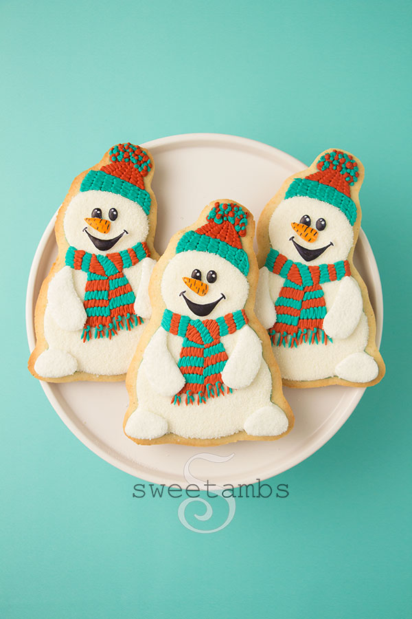 3 snowman cookies on a white plate on a teal background. The snowman cookies are decorated with teal and red scarves and hats. They have carrot noses and cartoonish eyes and a mouth.