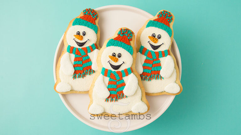 3 snowman cookies on a white plate on a teal background. The snowman cookies are decorated with teal and red scarves and hats. They have carrot noses and cartoonish eyes and a mouth.