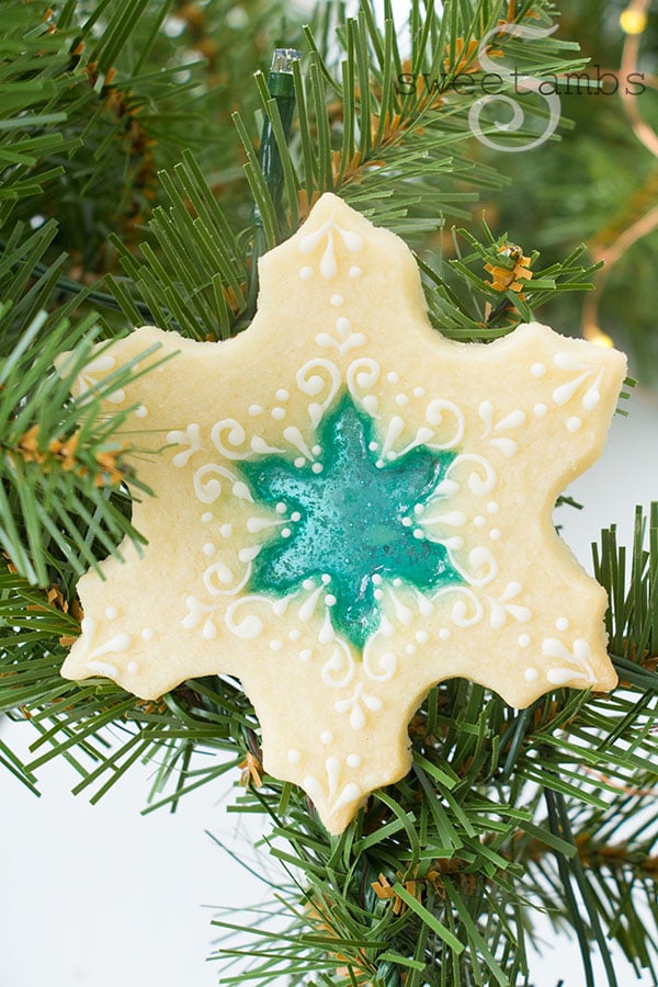 Beautiful stained glass cookies - snowflake cookies with a blue hard candy center that resembles stained glass. The cookies are mostly bare with a little bit of delicate royal icing decorations piped on them. The cookie is laying on sprigs of evergreen on a white background.