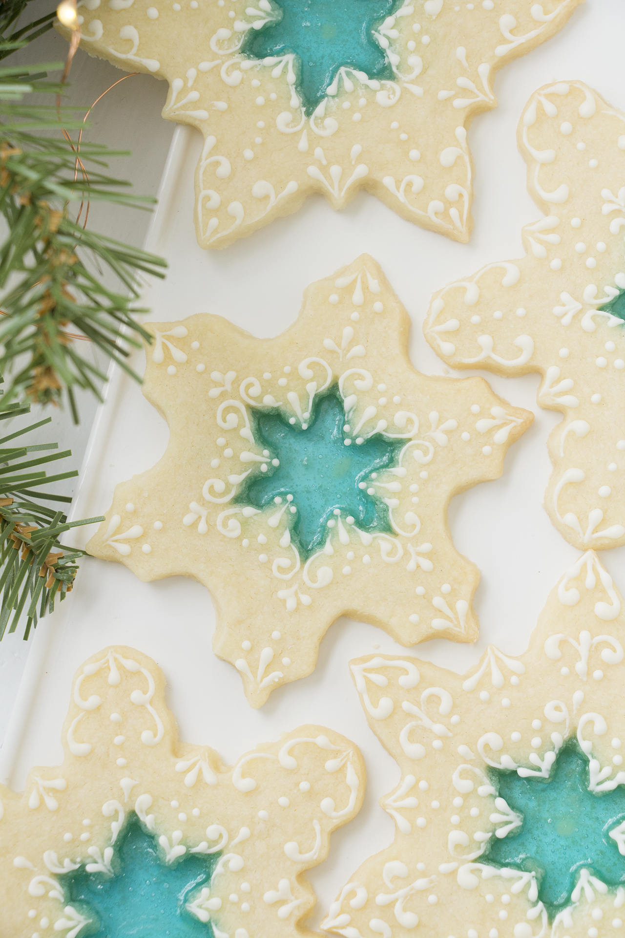 Beautiful stained glass cookies - snowflake cookies with a blue hard candy center that resembles stained glass. The cookies are mostly bare with a little bit of delicate royal icing decorations piped on them. The cookies are on a white rectangular platter on a white background. There is a sprig of evergreen in the foreground.