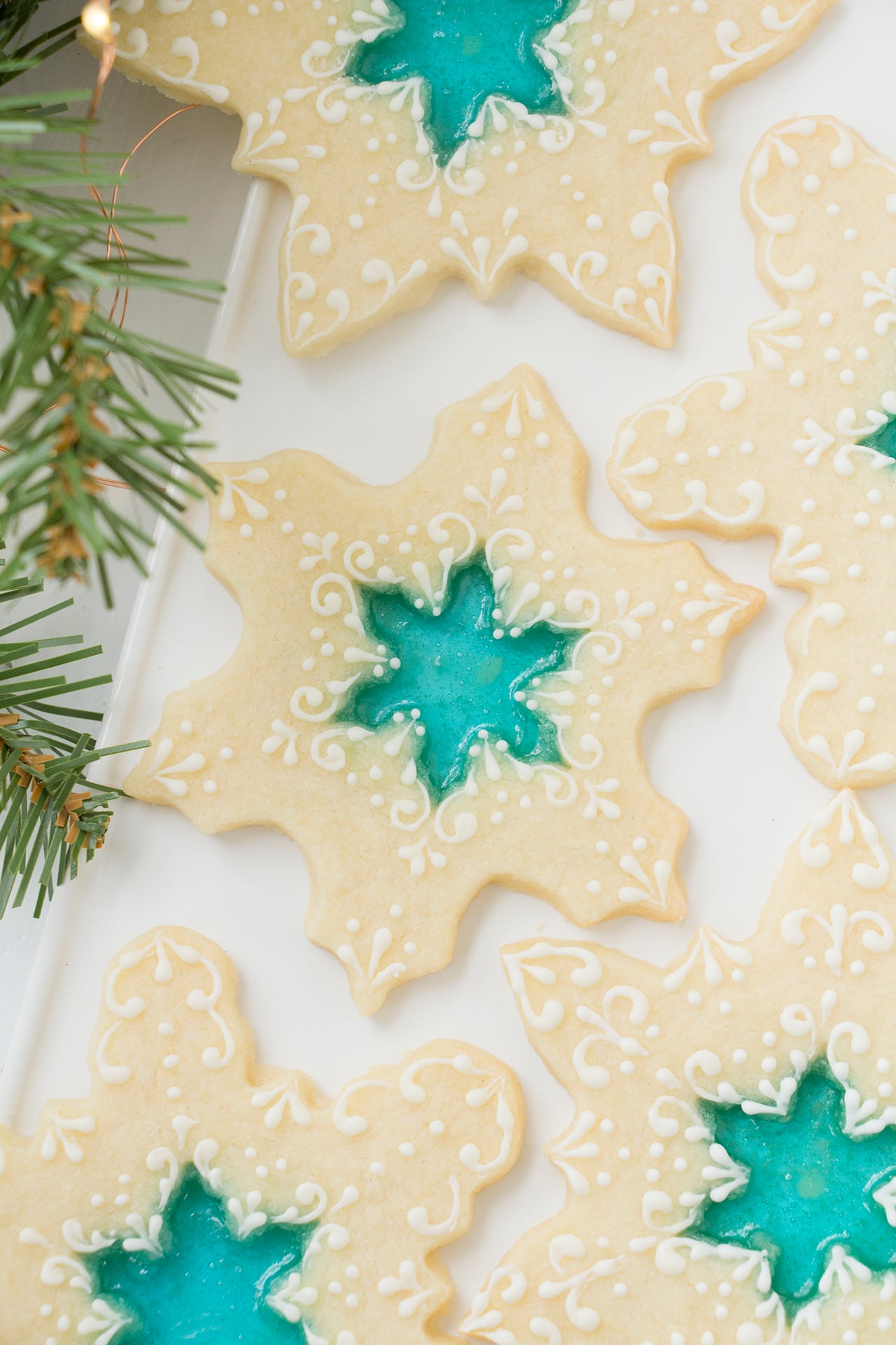 Beautiful stained glass cookies - snowflake cookies with a blue hard candy center that resembles stained glass. The cookies are mostly bare with a little bit of delicate royal icing decorations piped on them. The cookies are on a white rectangular platter on a white background. There is a sprig of evergreen in the foreground.