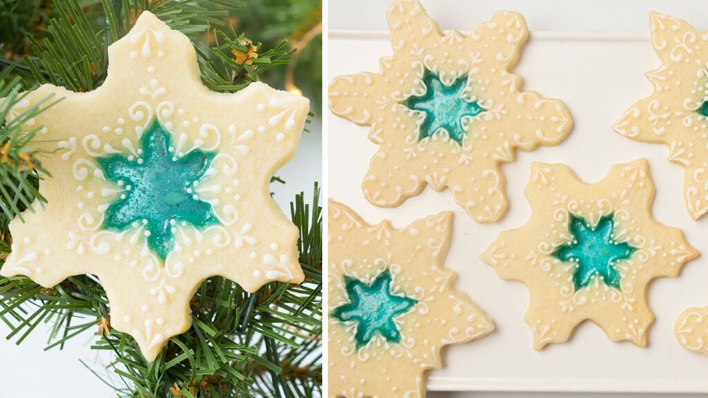 Beautiful stained glass cookies - snowflake cookies with a blue hard candy center that resembles stained glass. The cookies are mostly bare with a little bit of delicate royal icing decorations piped on them. The cookies are on a white rectangular platter on a white background. The cookie on the left side is laying on sprigs of evergreen.