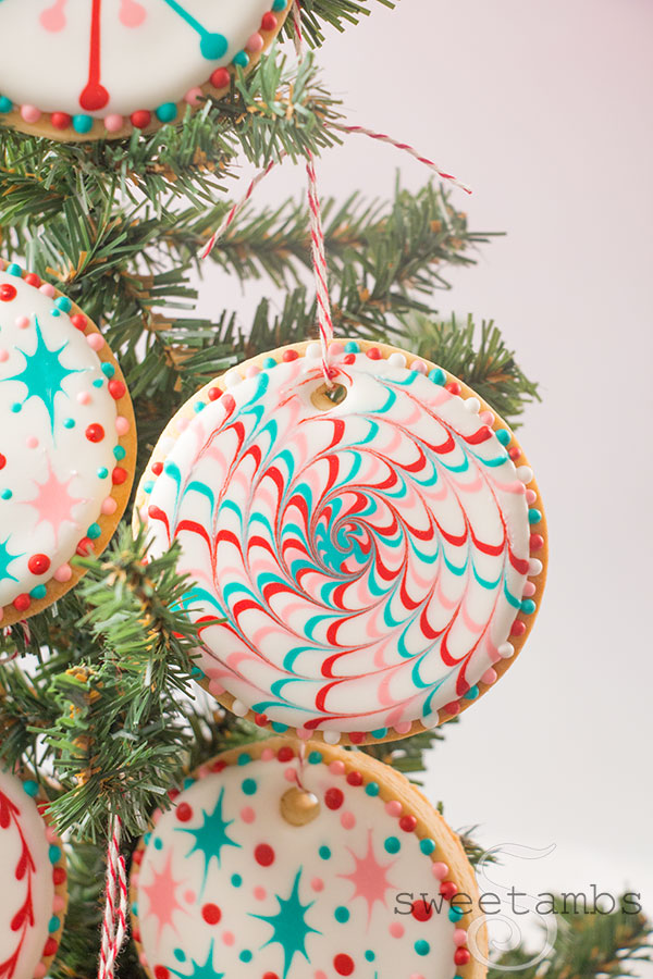 A closeup of Christmas Cookie Ornaments - Round cookies decorated with white, teal, red, and pink designs inspired by a 1960s Christmas. The cookies are hanging on a Christmas tree in front of a light pink background.