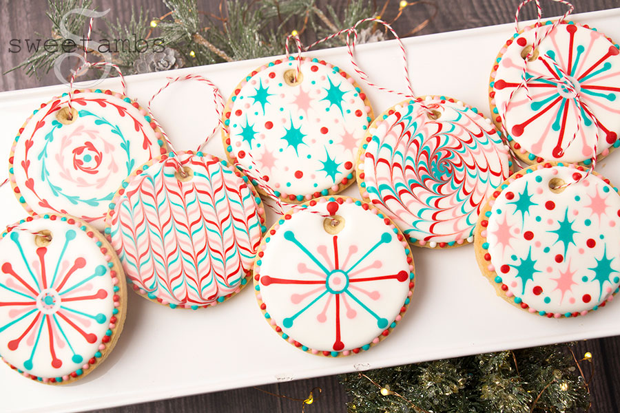 Christmas Cookie Ornaments - Round cookies decorated with white, teal, red, and pink designs inspired by a 1960s Christmas. The cookies have a string at the top so they can be hung on a tree. They're on a white rectangular platter on a wood background with some greenery and twinkle lights on the edges of the frame.