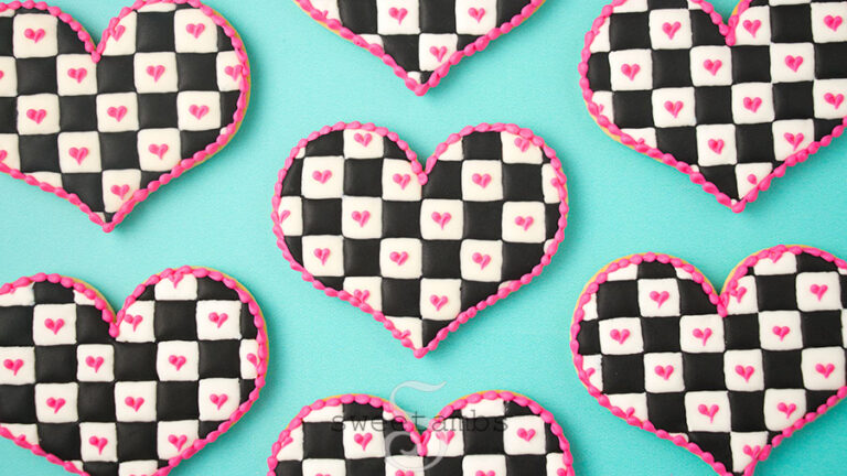 Checkerboard icing cookies - Heart shaped cookies decorated with a black and white checkered icing pattern with royal icing. There are pink hearts on the white squares and a pink bead border around the edge of the cookies. The cookies are on a teal background.