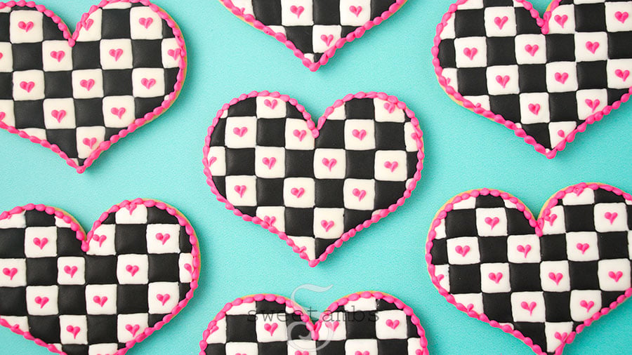 Checkerboard icing cookies - Heart shaped cookies decorated with a black and white checkered icing pattern with royal icing. There are pink hearts on the white squares and a pink bead border around the edge of the cookies. The cookies are on a teal background.