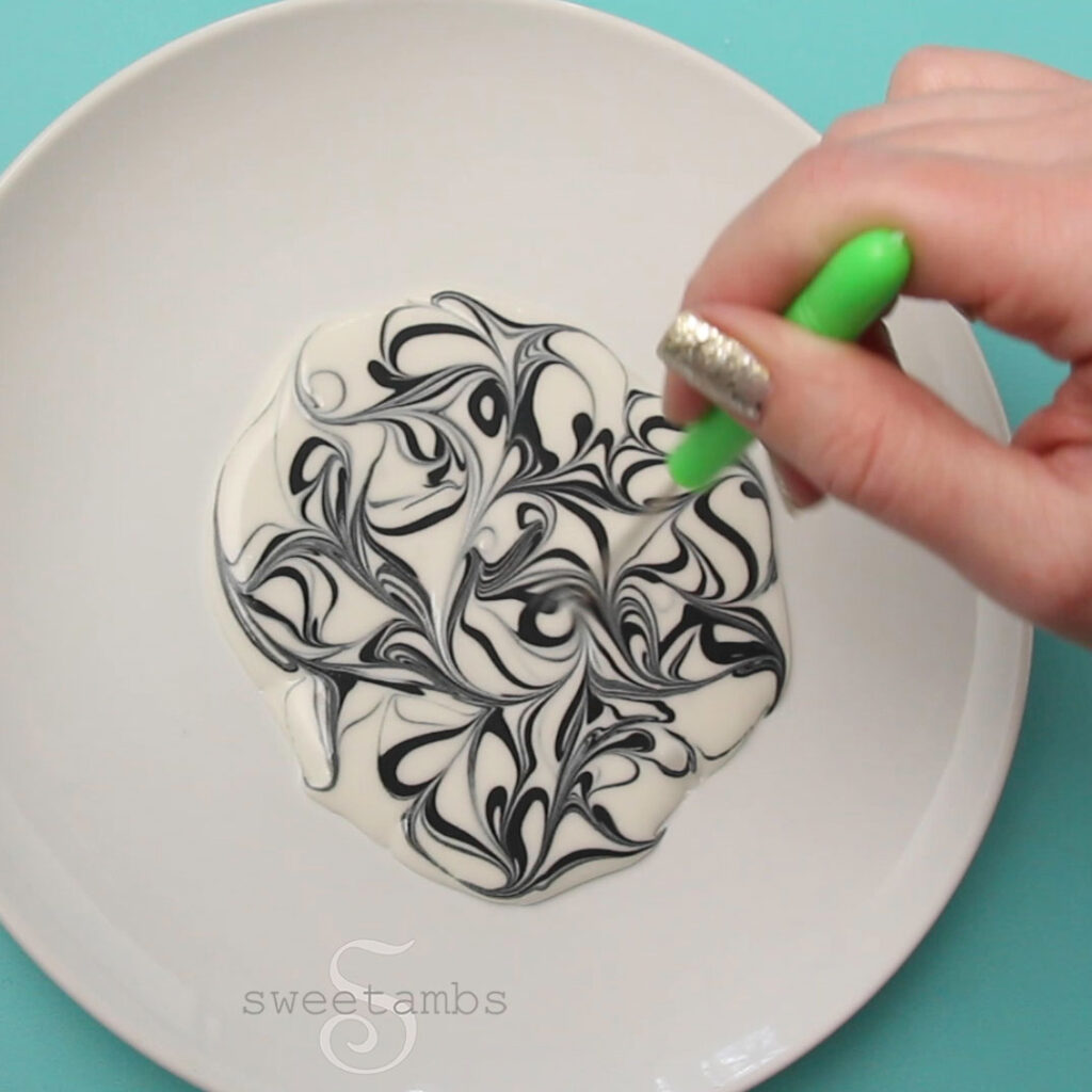 A dish of black and white royal icing being swirled with a scribe tool. The hand holding the scribe tool has silver glitter nail polish. The dish of icing is on a teal background.