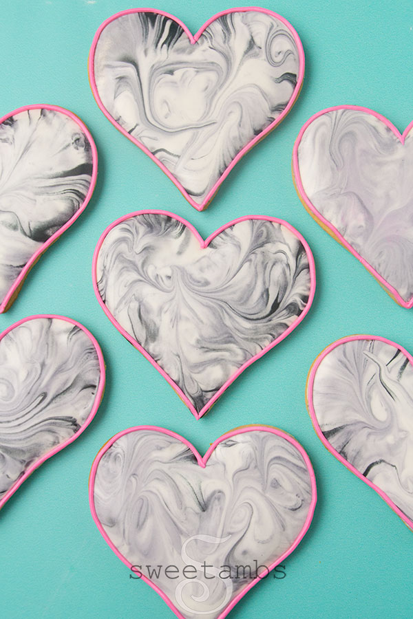 Heart shaped cookies decorated with black and white marble royal icing. The cookies have a think pink outline. They're on a teal background.