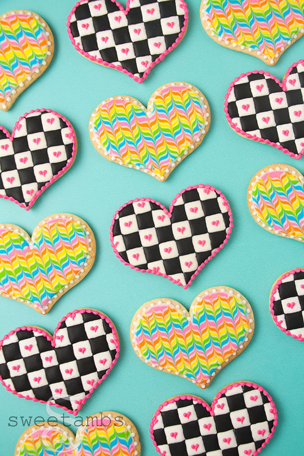 Checkerboard icing cookies and rainbow herringbone cookies - Heart shaped cookies decorated with a black and white checkered icing pattern with royal icing. There are pink hearts on the white squares and a pink bead border around the edge of the cookies. In between the checkerboard cookies are heart cookies decorated with a rainbow herringbone pattern. The cookies are on a teal background.