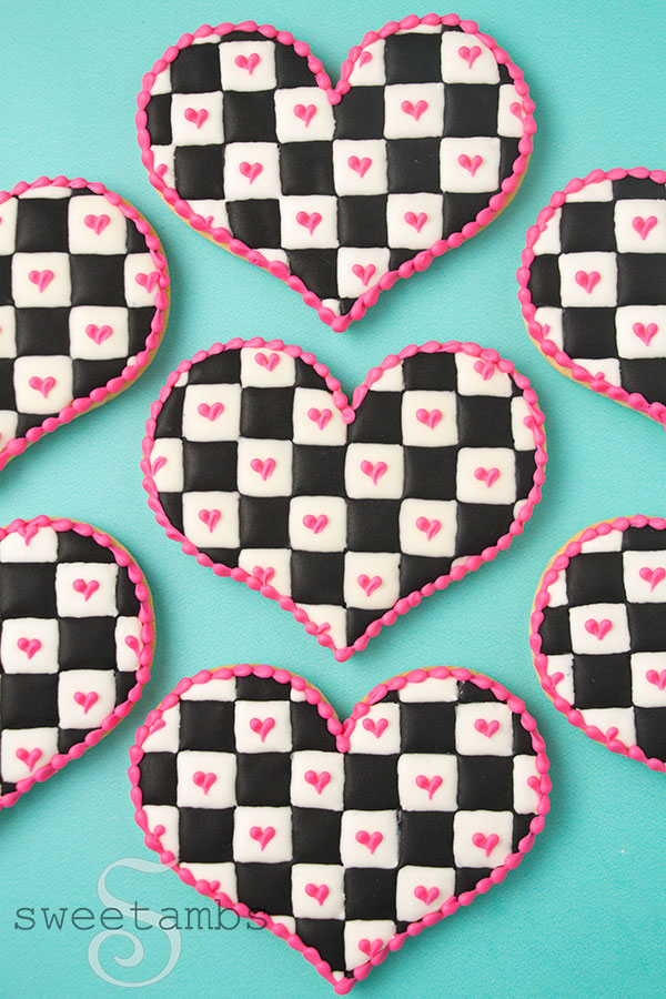 Checkerboard icing cookies - Heart shaped cookies decorated with a black and white checkered icing pattern with royal icing. There are pink hearts on the white squares and a pink bead border around the edge of the cookies. The cookies are on a teal background.