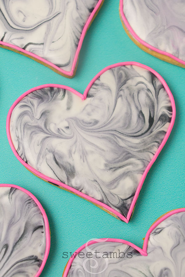close up of Heart shaped cookies decorated with black and white marble royal icing. The cookies have a think pink outline. They're on a teal background.
