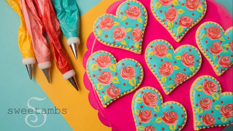 Valentine's Day decorated cookies - heart shaped cookies decorated with wet-on-wet royal icing roses. The base layer of icing is teal and the roses are bright pink. There are red dots and yellow leaves on the cookies as well as a pink and yellow dot border around the edges. The cookies are on a bright pink scalloped cake plate on a golden yellow and teal background. There are piping bags filled with icing next to the cookies.