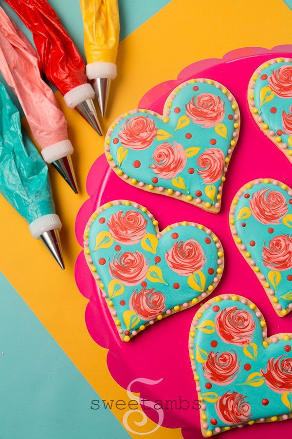 Valentine's Day decorated cookies - heart shaped cookies decorated with wet-on-wet royal icing roses. The base layer of icing is teal and the roses are bright pink. There are red dots and yellow leaves on the cookies as well as a pink and yellow dot border around the edges. The cookies are on a bright pink scalloped cake plate on a golden yellow and teal background. There are piping bags filled with icing next to the cookies.