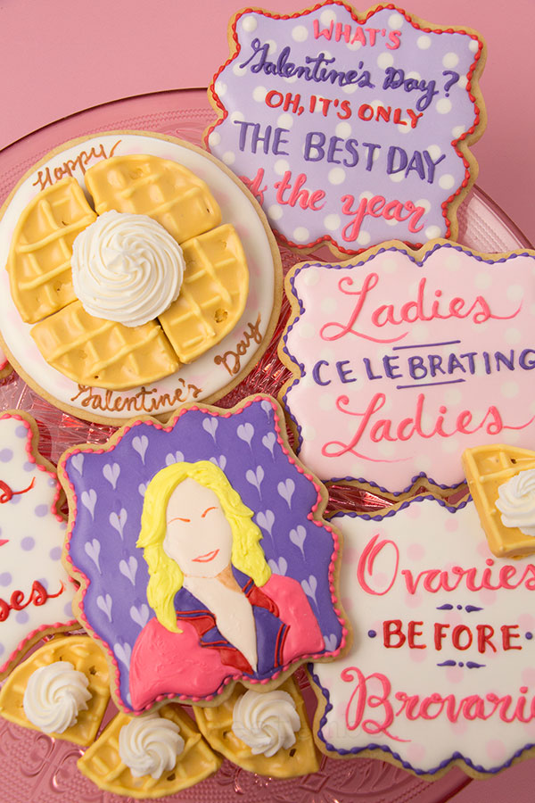 A set of cookies celebrating Galentine's Day from Parks and Rec. The set includes plaque shaped cookies decorated in white, pink and purple icing with quotes from the Galentine's Day episode. There is a plaque shaped cookie in the center decorated with purple hearts and a cartoonish illustration of Leslie Knope done in royal icing. Above that is a cookie decorated to look like a waffle on a plate topped with whipped cream. There are waffle cookies scattered around the platter. The cookies are on a pink cake stand on a pink background.