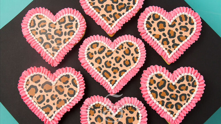 Leopard print cookies for Valentine's Day - A set of 7 heart shaped cookies decorated with a brown and black leopard print pattern in royal icing. There is a bright pink ruffle border around each cookie as well as a white bead border on the inside edge of the ruffle. The cookies are sitting on a black diamond shaped background over a teal background.