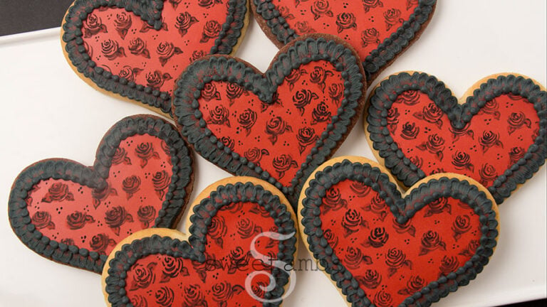 Painting on cookies - A set of heart shaped cookies decorated with red royal icing and hand painted black roses. The cookies have a black brush embroidered and beaded border around the edges. They are on a white rectangular platter on a black background.