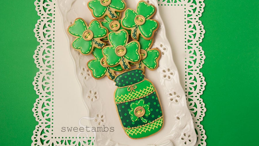 Saint patrick's day cookies - a set of cookies decorated to look like a bouquet of shamrocks in a mason jar. The cookies are decorated with royal icing in different shades of green. There are polka dots, lace, stitches, and gold buttons on the cookies to make them look as if they are quilted. The cookies are on a rectangular platter with eyelets and the platter is on a square of white paper with a lace edge over a green background.