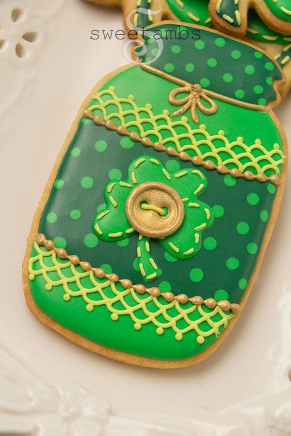 A closeup of Saint Patrick's Day Cookies - this cookie is shaped like a mason jar and decorated in royal icing. It is decorated in 3 shades of green with polka dots, lace, stitches, and a gold button.
