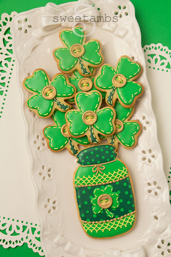 Saint patrick's day cookies - a set of cookies decorated to look like a bouquet of shamrocks in a mason jar. The cookies are decorated with royal icing in different shades of green. There are polka dots, lace, stitches, and gold buttons on the cookies to make them look as if they are quilted. The cookies are on a rectangular platter with eyelets and the platter is on a square of white paper with a lace edge over a green background.