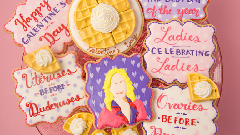 A set of cookies celebrating Galentine's Day from Parks and Rec. The set includes plaque shaped cookies decorated in white, pink and purple icing with quotes from the Galentine's Day episode. There is a plaque shaped cookie in the center decorated with purple hearts and a cartoonish illustration of Leslie Knope done in royal icing. Above that is a cookie decorated to look like a waffle on a plate topped with whipped cream. There are waffle cookies scattered around the platter. The cookies are on a pink cake stand on a pink background.