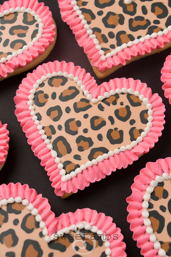 Leopard print cookies for Valentine's Day - A set of heart shaped cookies decorated with a brown and black leopard print pattern in royal icing. There is a bright pink ruffle border around each cookie as well as a white bead border on the inside edge of the ruffle. The cookies are sitting on a black background.