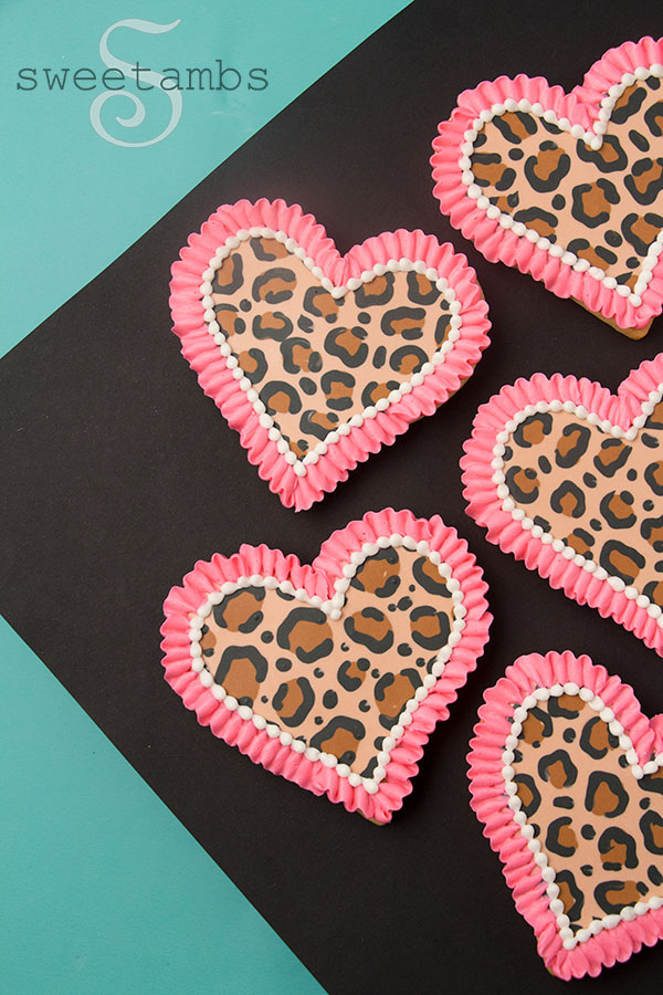 Leopard print cookies for Valentine's Day - A set of heart shaped cookies decorated with a brown and black leopard print pattern in royal icing. There is a bright pink ruffle border around each cookie as well as a white bead border on the inside edge of the ruffle. The cookies are sitting on a black diamond shaped background over a teal background.