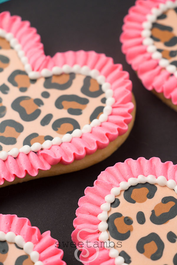 Close up of Leopard print cookies for Valentine's Day - A set of heart shaped cookies decorated with a brown and black leopard print pattern in royal icing. There is a bright pink ruffle border around each cookie as well as a white bead border on the inside edge of the ruffle. The cookies are sitting on a black background.
