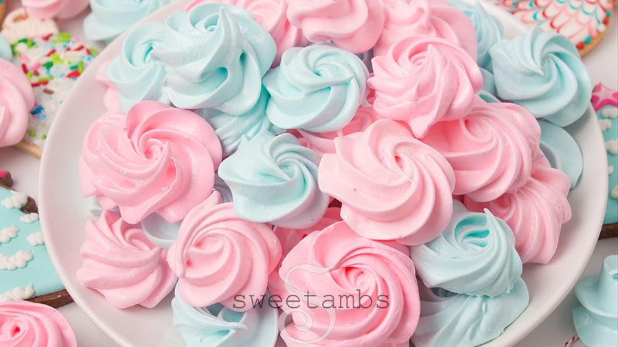 Pink and blue meringue cookies on a round white plate. There are meringue cookies scattered around the plate along with link and blue decorated cookies.