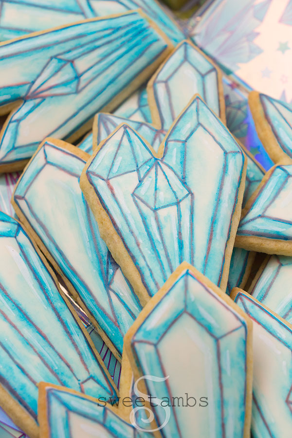 Watercolor crystal cookies - Cookies decorated with royal icing and watercolor technique to look like crystals. There are outlines on the cookies drawn with edible ink marker. The cookies are on a holographic background with a crystal design.