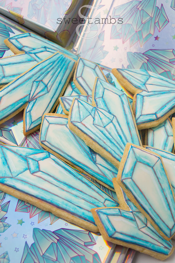 Watercolor crystal cookies - Cookies decorated with royal icing and watercolor technique to look like crystals. There are outlines on the cookies drawn with edible ink marker. The cookies are on a holographic background with a crystal design.