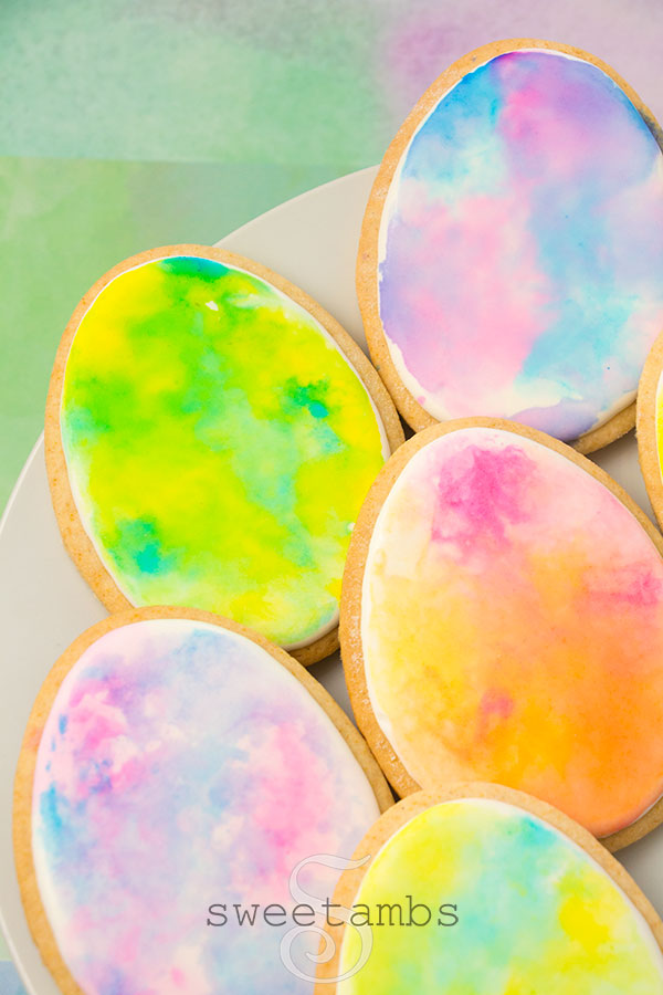watercolor easter egg cookies - Egg shaped cookies decorated with royal icing and watercolor in pastel colors. The cookies are on a white plate over a watercolor paper background.