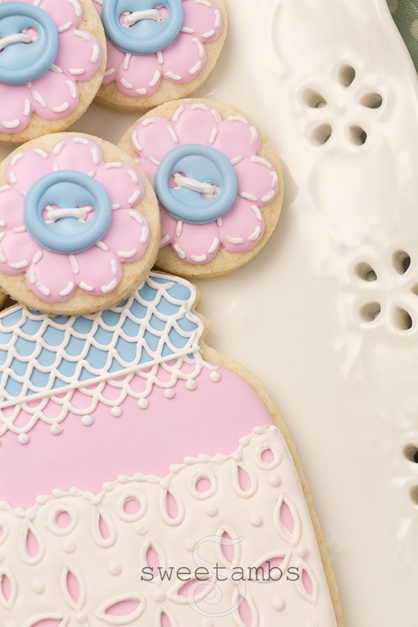 A close up of a decorated mason jar cookie with a pink base and a blue lid. The mason jar cookie has a band of royal icing eyelet lace across the middle and the lid is covered with royal icing lace. There are several small cookies on top of the jar to look like a bouquet. The smaller cookies are decorated to look like flowers with royal icing stitches and a royal icing button in the center of each flower. The cookies are on a porcelain eyelet lace platter.