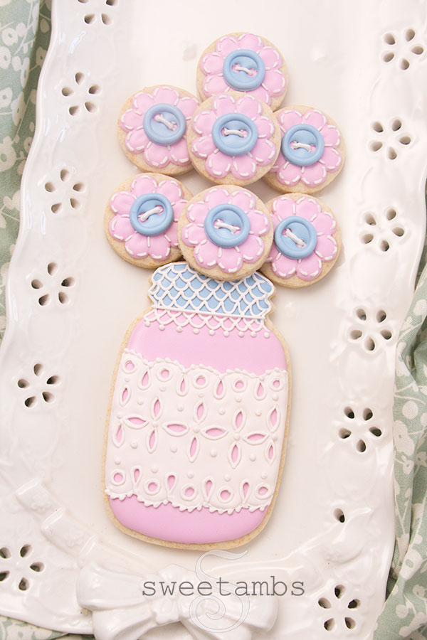 A decorated mason jar cookie with a pink base and a blue lid. The mason jar cookie has a band of royal icing eyelet lace across the middle and the lid is covered with royal icing lace. There are several small cookies on top of the jar to look like a bouquet. The smaller cookies are decorated to look like flowers with royal icing stitches and a royal icing button in the center of each flower. The cookies are on a porcelain eyelet lace platter on a piece of green and white floral cloth.