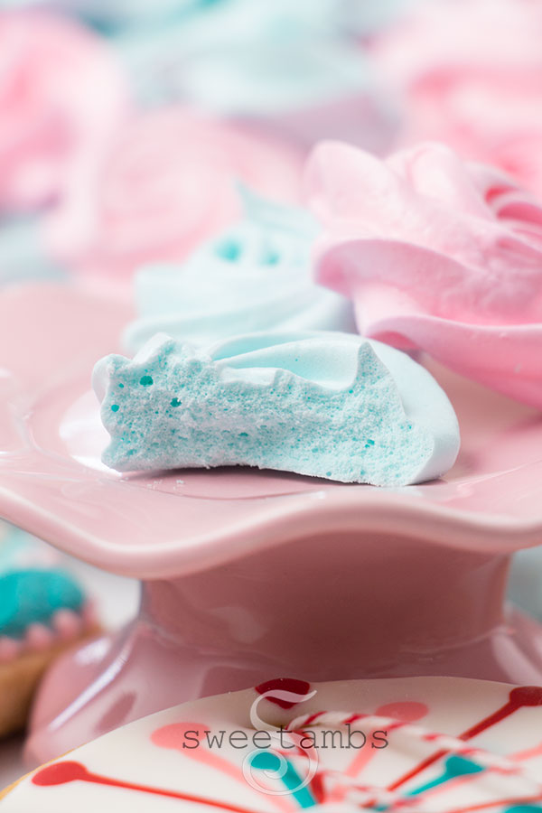 A blue meringue cookie with a bite taken out and showing the inside of the meringue. There  are two meringue cookies on the plate and more meringue cookies in the background. The plate is a small pink cake plate with a wavy edge. There is a brightly colored decorated cookie below the cake plate.