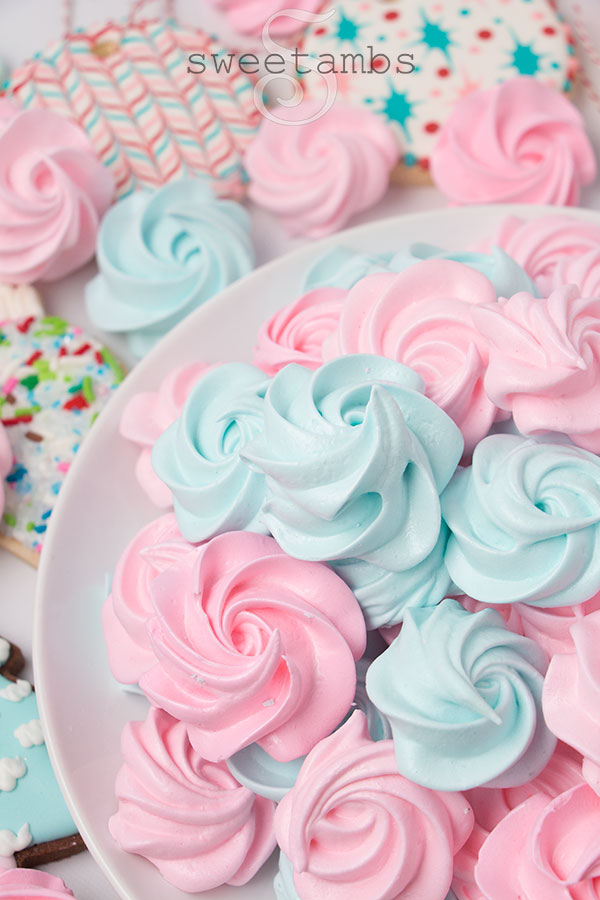 Pink and blue meringue cookies shaped like rosettes on a round white plate. There are meringue cookies scattered around the plate along with link and blue decorated cookies.