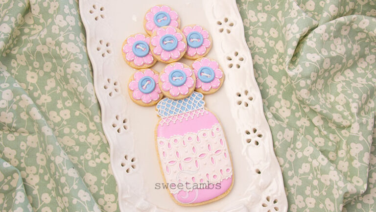 A decorated mason jar cookie with a pink base and a blue lid. The mason jar cookie has a band of royal icing eyelet lace across the middle and the lid is covered with royal icing lace. There are several small cookies on top of the jar to look like a bouquet. The smaller cookies are decorated to look like flowers with royal icing stitches and a royal icing button in the center of each flower. The cookies are on a porcelain eyelet lace platter on a piece of green and white floral cloth.