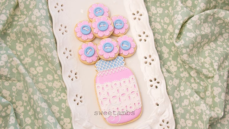 A decorated mason jar cookie with a pink base and a blue lid. The mason jar cookie has a band of royal icing eyelet lace across the middle and the lid is covered with royal icing lace. There are several small cookies on top of the jar to look like a bouquet. The smaller cookies are decorated to look like flowers with royal icing stitches and a royal icing button in the center of each flower. The cookies are on a porcelain eyelet lace platter on a piece of green and white floral cloth.